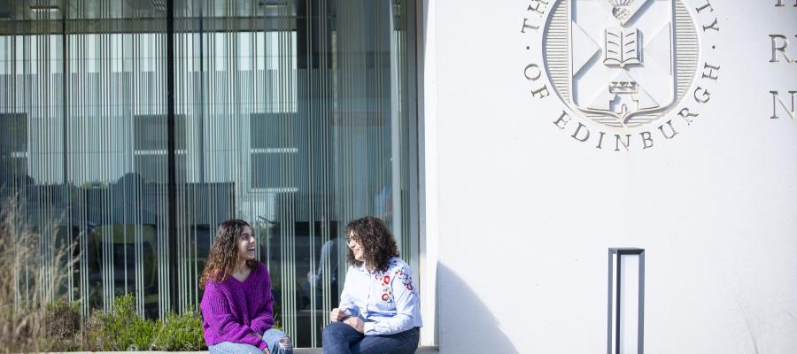 Two female students sitting outside a building, beside a University of Edinburgh logo on the wall