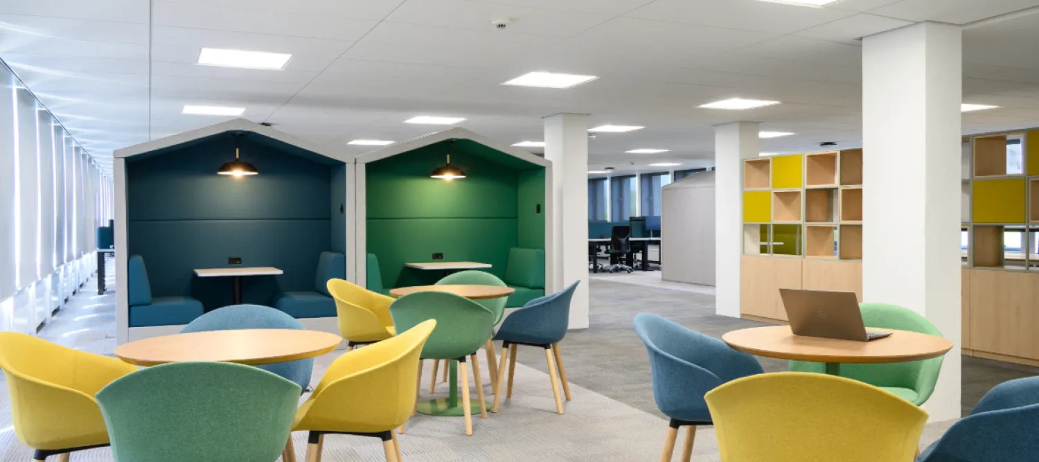 Office space with round three tables each with four chairs around them. Two meeting pods in the background