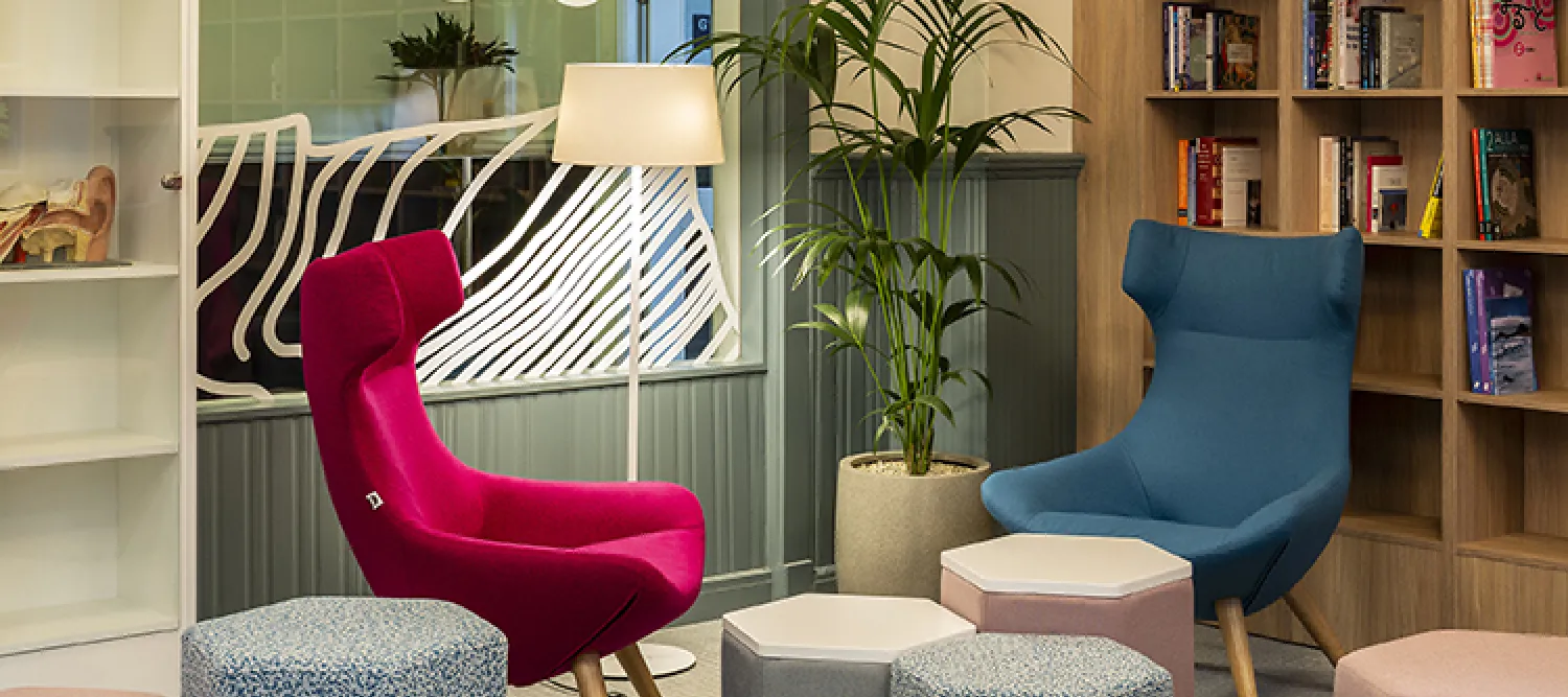 A room with one pink high back chair and one blue high back chair, grey and pink stools and book shelves