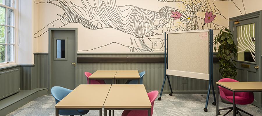 A room with tables and chairs, a pinboard on wheels, one wall has a large pattern of pink and white flowers