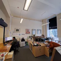 An office space with a person working on a computer and a person looking in a cupboard