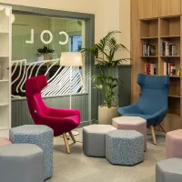 A room with one pink high back chair and one blue high back chair, grey and pink stools and book shelves