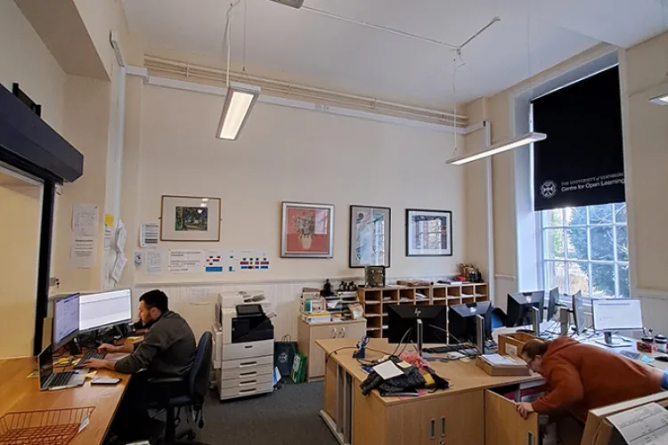 An office space with a person working on a computer and a person looking in a cupboard