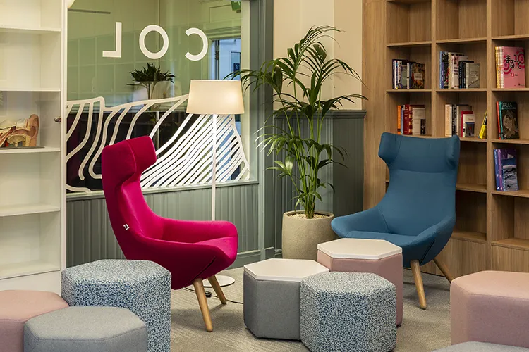 A room with one pink high back chair and one blue high back chair, grey and pink stools and book shelves