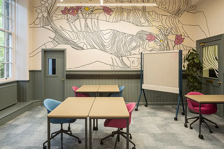 A room with tables and chairs, a pinboard on wheels, one wall has a large pattern of pink and white flowers